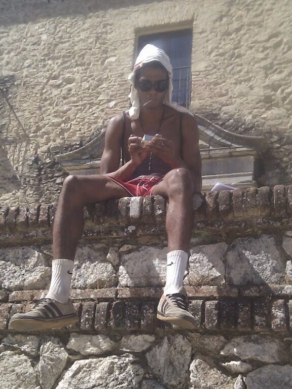 Manu at 63 kg, very skinny, sitting on a stone wall in a beanie and sunglasses