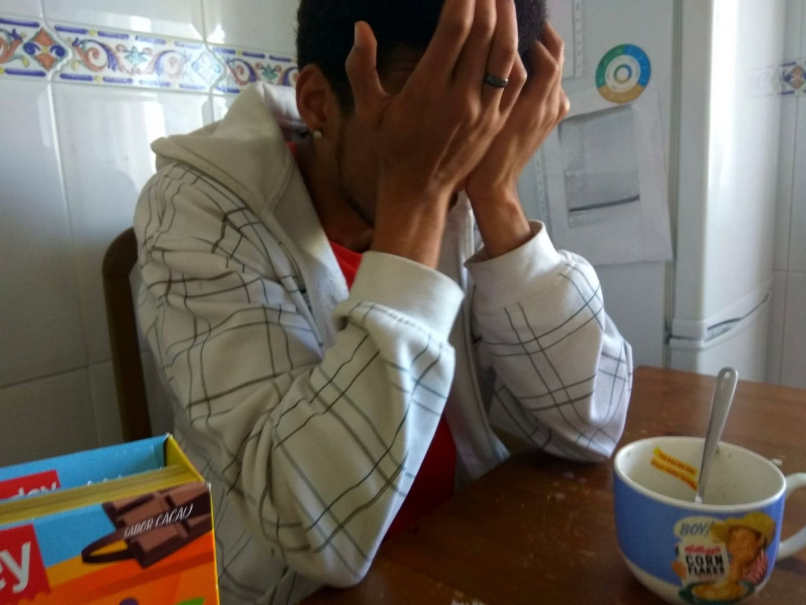 Manu sitting in the kitchen with his face in his hands and a bowl of cereal in front of him, frustrated