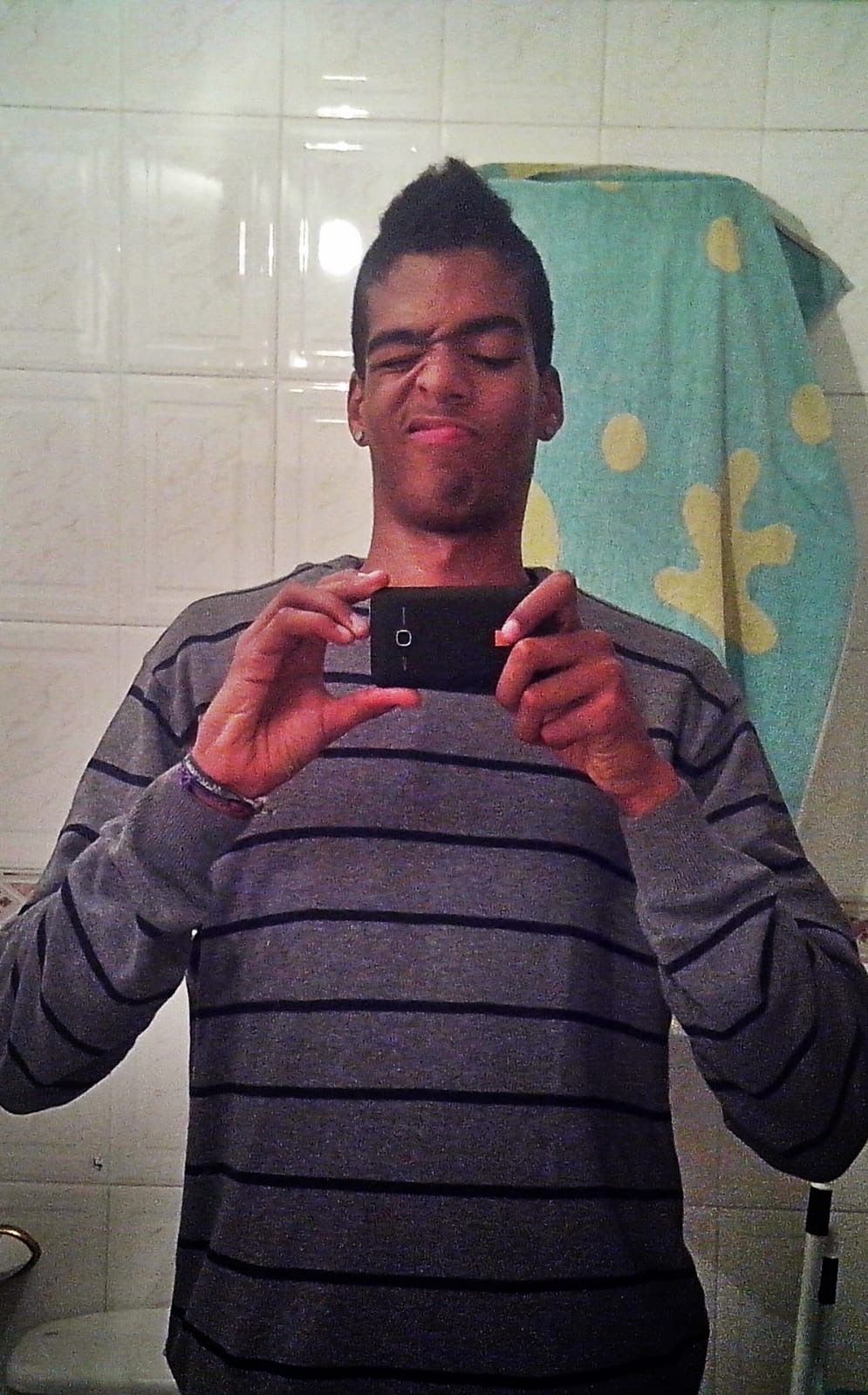 Manu early on, very skinny, in a striped sweater, taking a bathroom mirror selfie and pulling a face